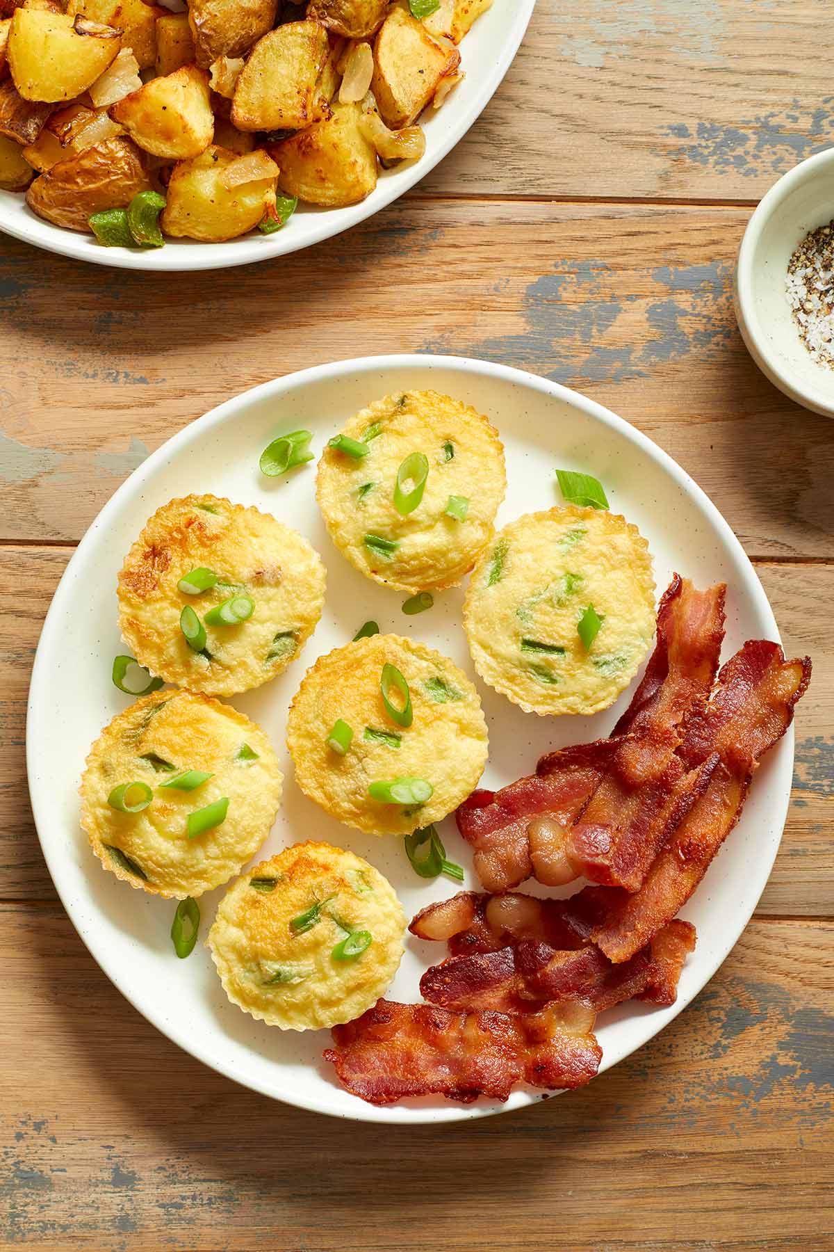 Egg muffins on a plate with slices of bacon and a plate of home fries on the side.