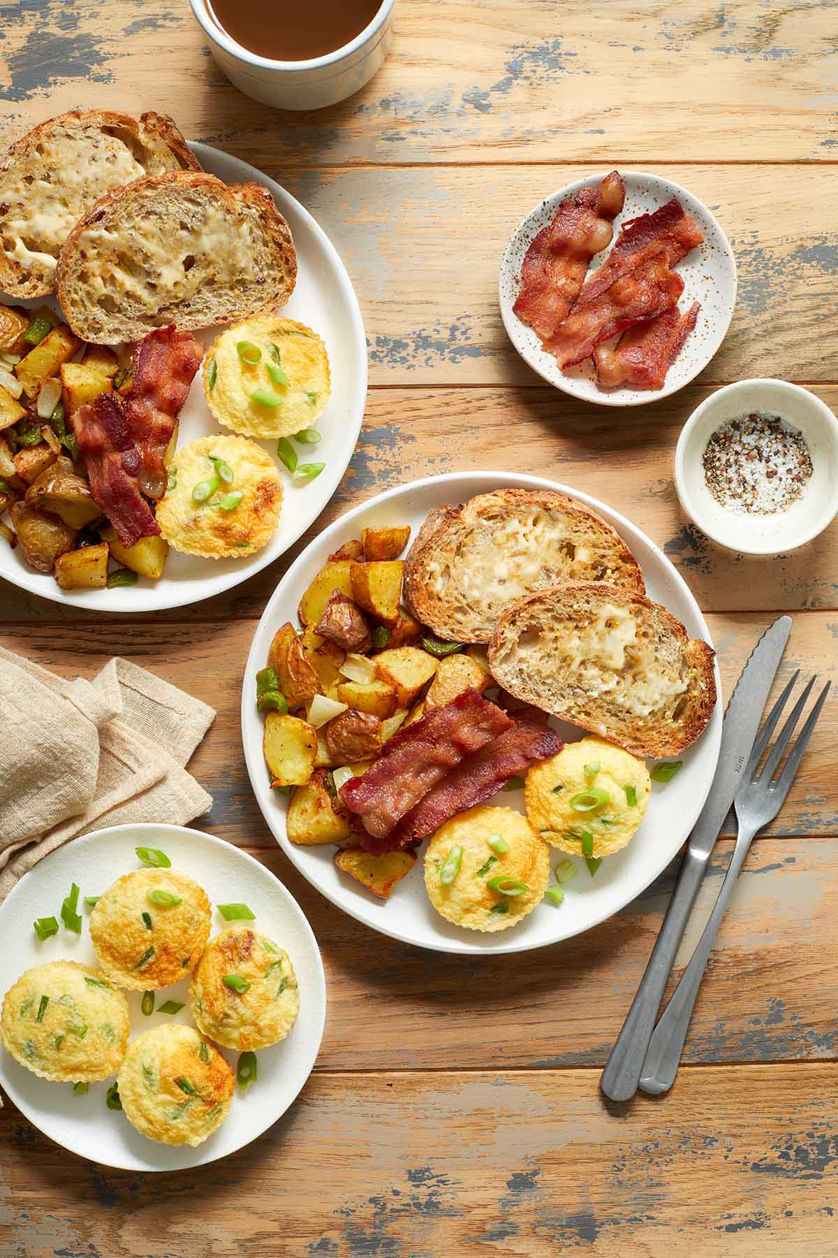 A breakfast setting with plates of egg bites, bacon, home fries and toast on a wooden table.