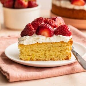 Side view of a slice of lemon almond flour cake on a plate with whipped cream and strawberries on top.