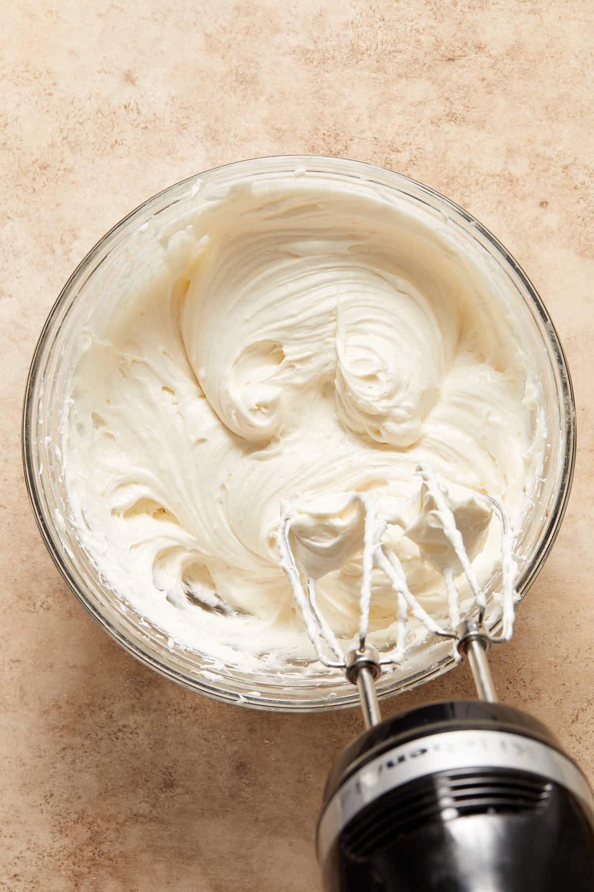 Overhead view of dairy-free cream cheese frosting in a glass bowl with a handheld electric mixer on the side.