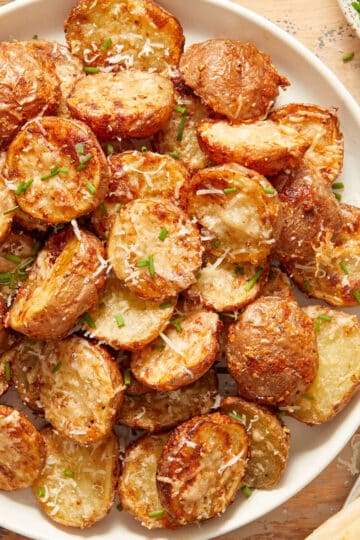 Overhead view of air fryer parmesan potatoes on a white plate next to chopped chives and parmesan cheese.