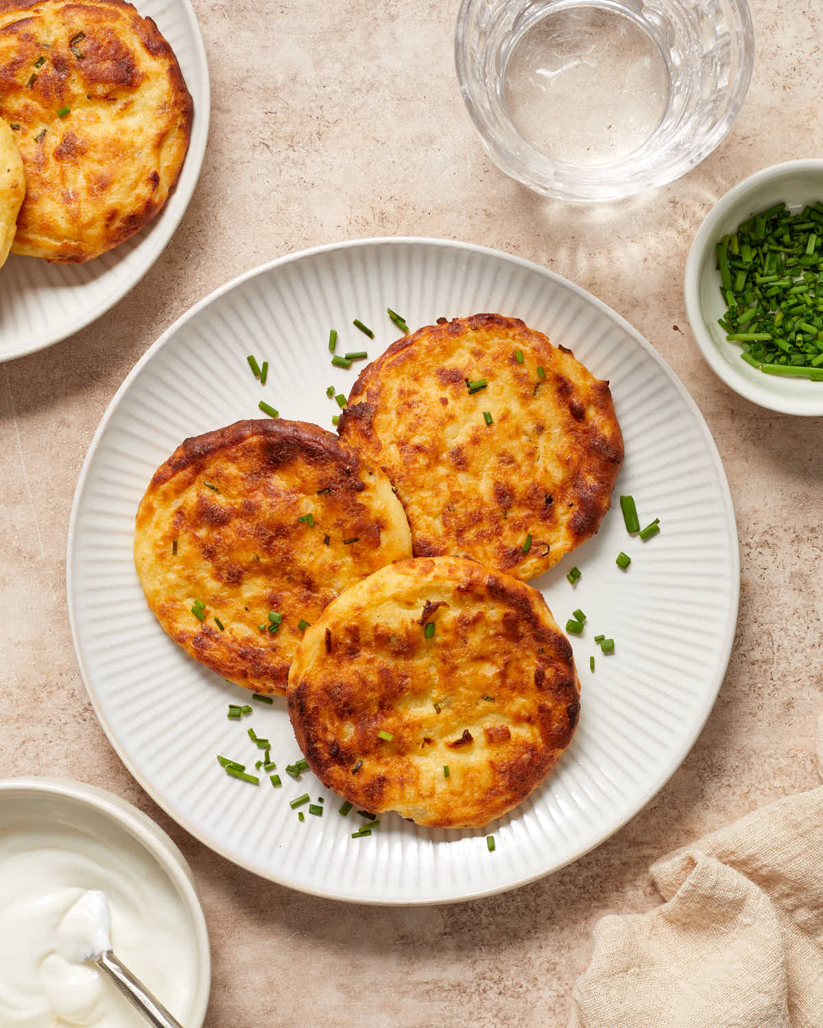 Three potato cakes on a plate next to some sour cream, chives and a glass of water.