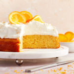 Side view of almond flour orange cake on a white cake stand with whipped cream and oranges on top and some slices of cake removed.