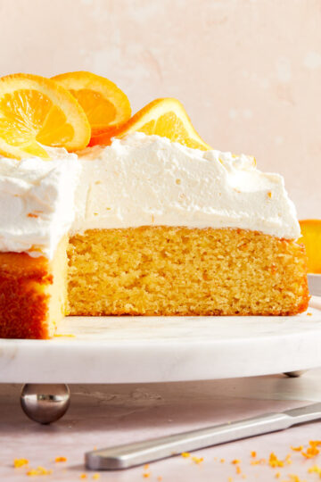 Side view of almond flour orange cake on a white cake stand with whipped cream and oranges on top and some slices of cake removed.