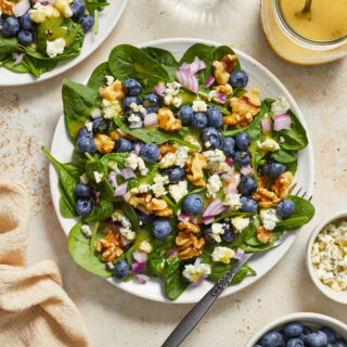 Blueberry spinach salad on a white plate with walnuts, red onion, blue cheese, blueberries and dijon dressing on top.