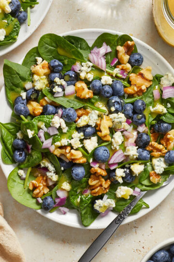 Blueberry spinach salad on a white plate with walnuts, red onion, blue cheese, blueberries and dijon dressing on top.