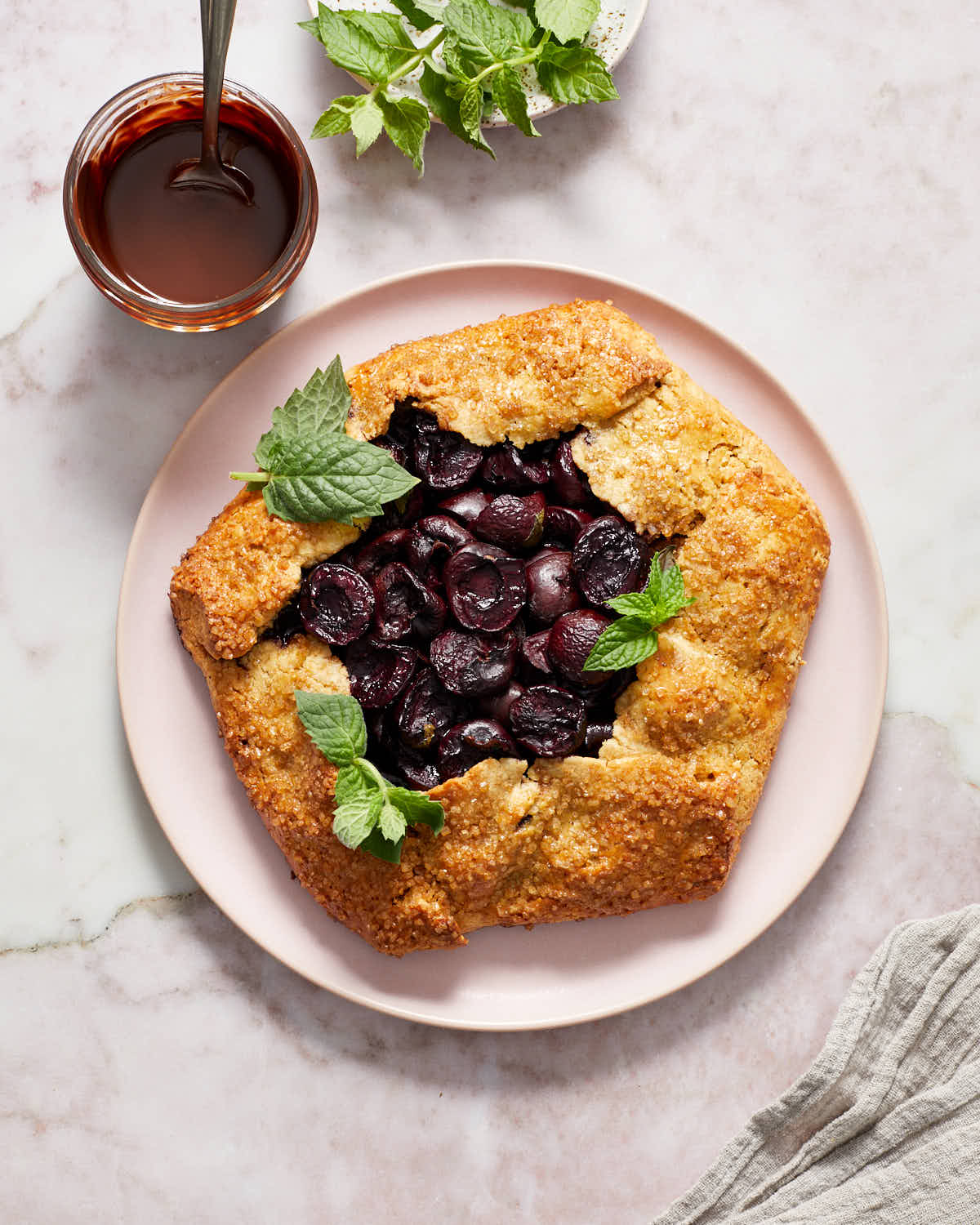 Baked cherry galette on a pink plate with melted chocolate and fresh mint leaves next to it.