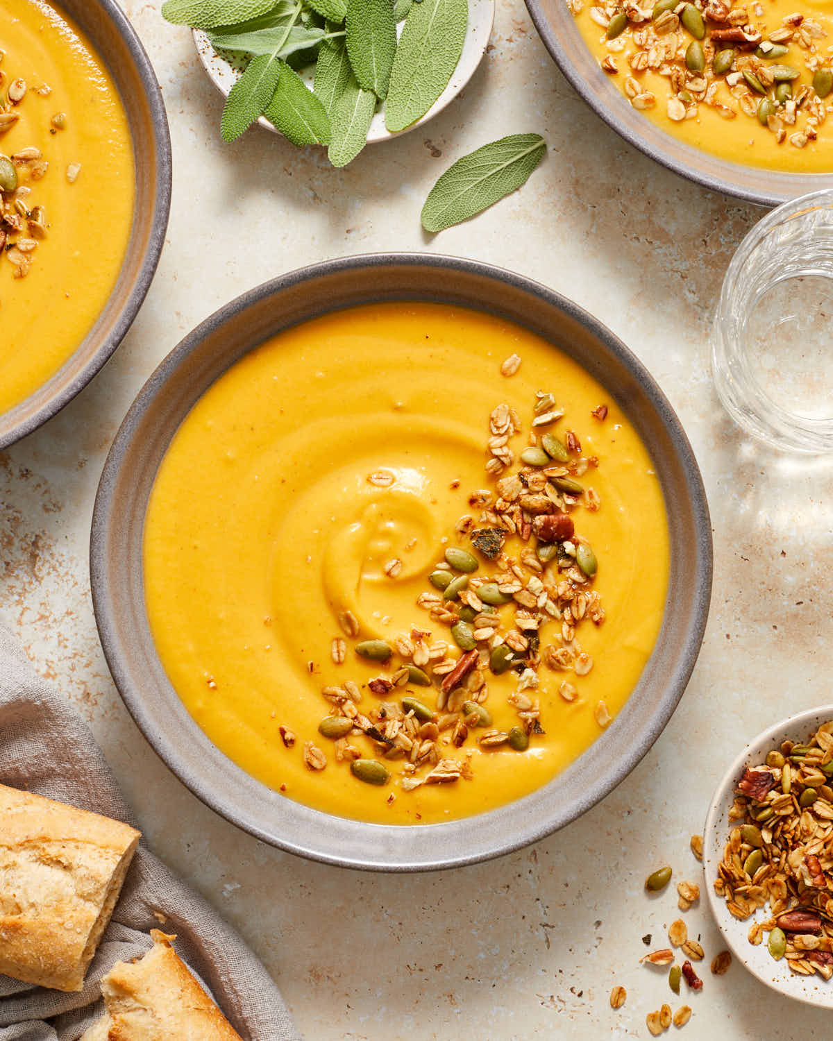 Bowls of squash and carrot soup next to bread, granola and sage.