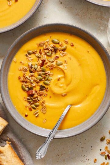 A bowl of carrot and butternut squash soup topped with savory granola next to some crusty bread.