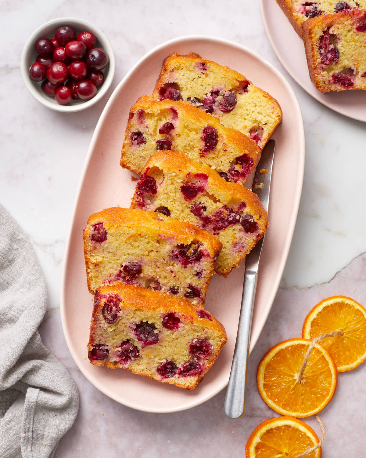 Slices of cranberry orange bread arranged on a pink platter with cranberries and orange slices to the side.