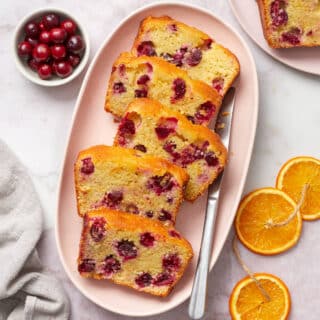 Slices of almond flour cranberry orange bread on a pink oval platter with orange slices and cranberries on the side.