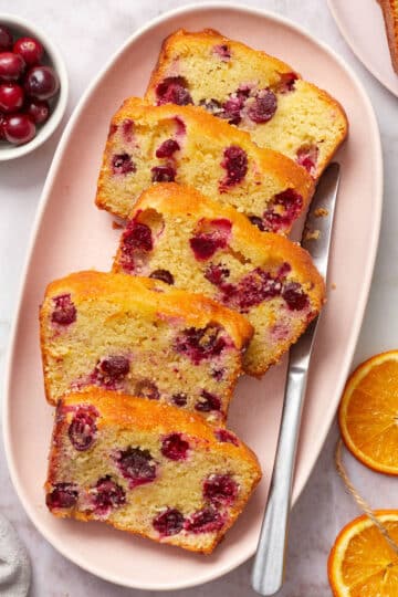 Slices of almond flour cranberry orange bread on a pink oval platter with orange slices and cranberries on the side.