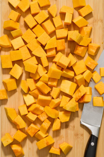 Peeled, cut and cubed butternut squash on a wooden cutting board with a chef's knife.