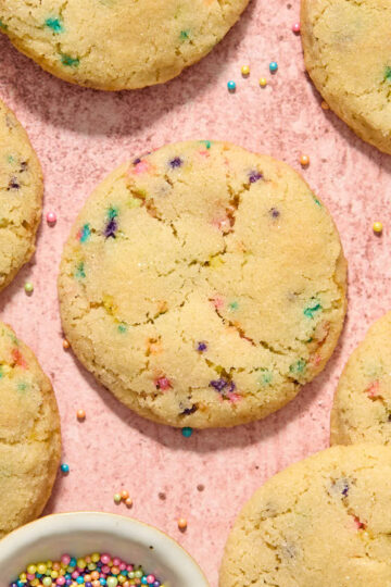 Almond flour sugar cookies (with sprinkles) arranged on a pink surface with a bowl of sprinkles on the side.