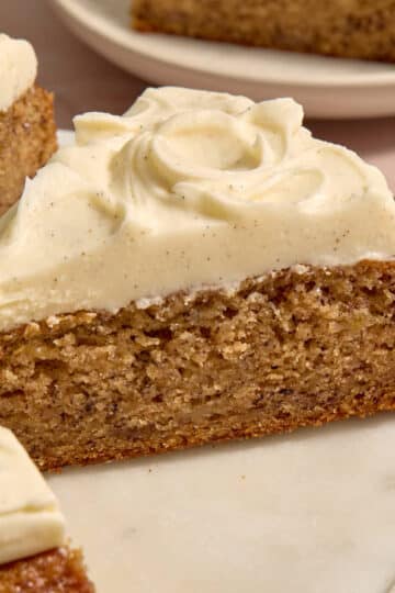A slice of eggless banana cake topped with cream cheese frosting being removed from the cake platter.