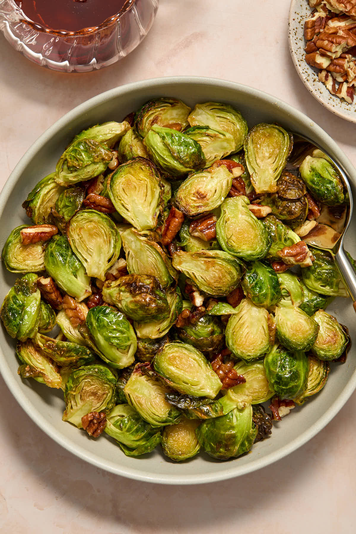 Brussels sprouts and pecans served in a grey bowl with chopped pecans and maple syrup on the side.