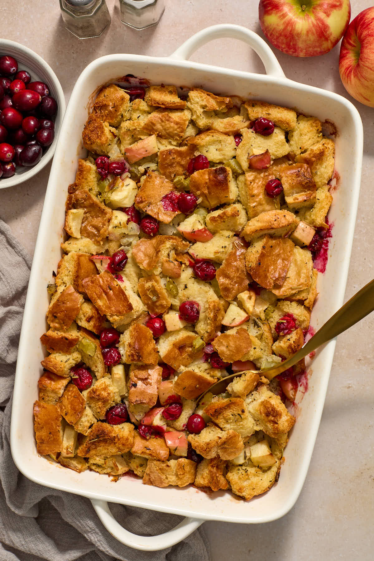 Stuffing in a white baking dish with a spoon inserted into it, and cranberries and apples on the side.