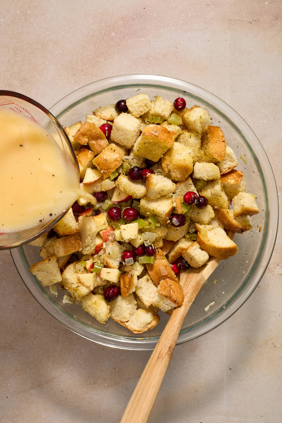 Stuffing ingredients mixed together in a glass bowl with a wooden spoon and egg/broth mixture being poured over it.