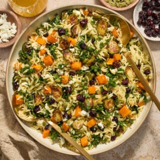 Fall orzo salad served in a large bowl with salad servers and extra vinaigrette and toppings on the side.