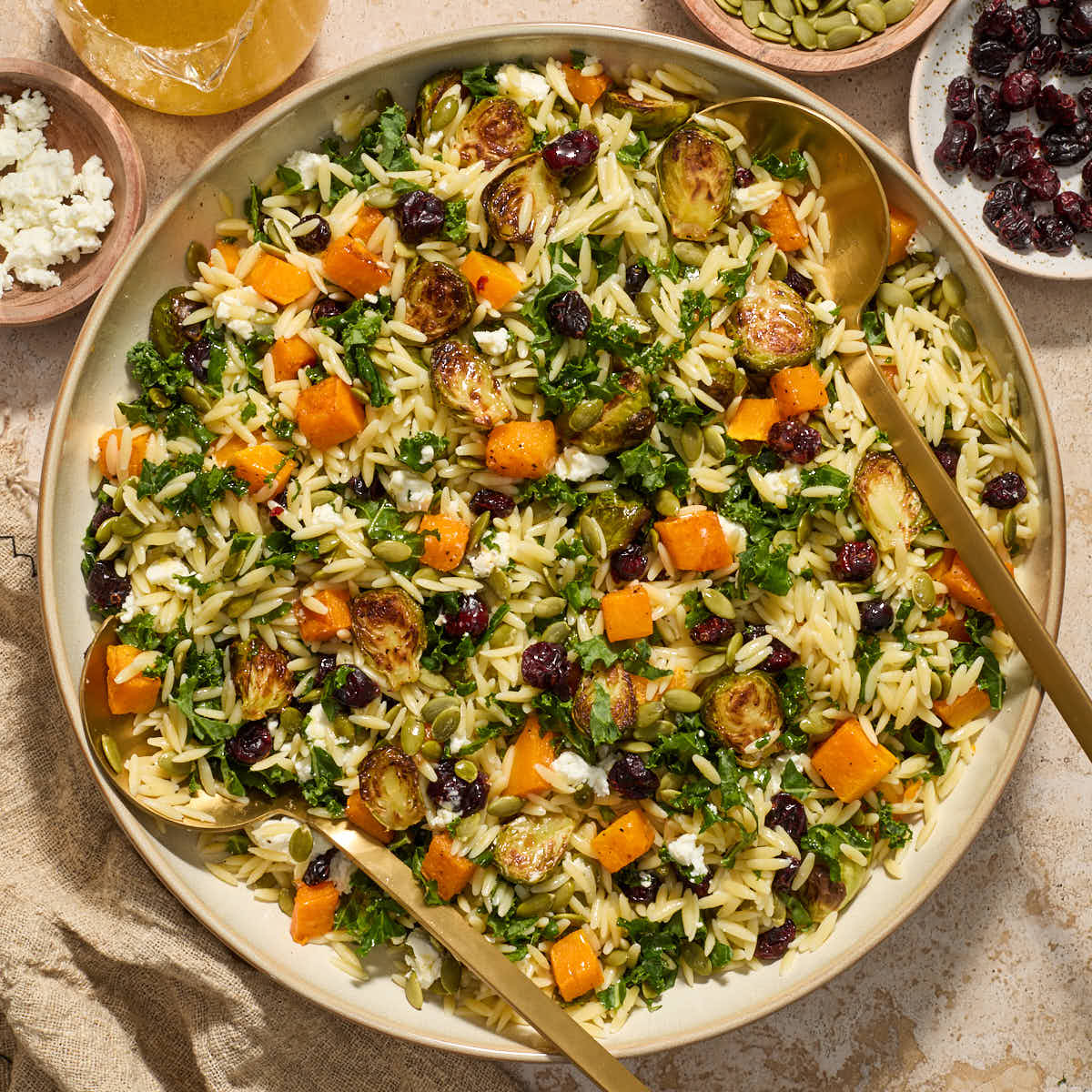Fall orzo salad served in a large bowl with salad servers and extra vinaigrette and toppings on the side.