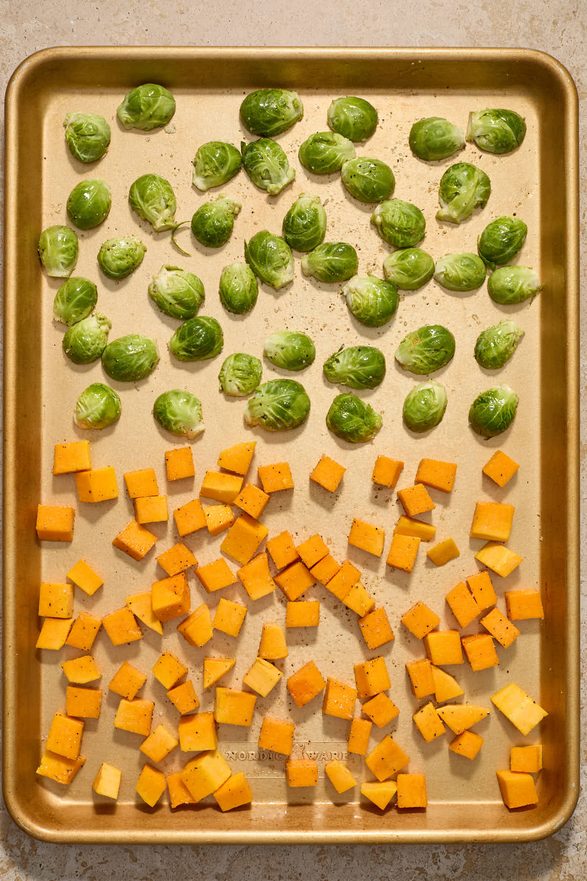 Cubed squash and halved Brussels sprouts seasoned and spread out on a sheet pan for roasting.