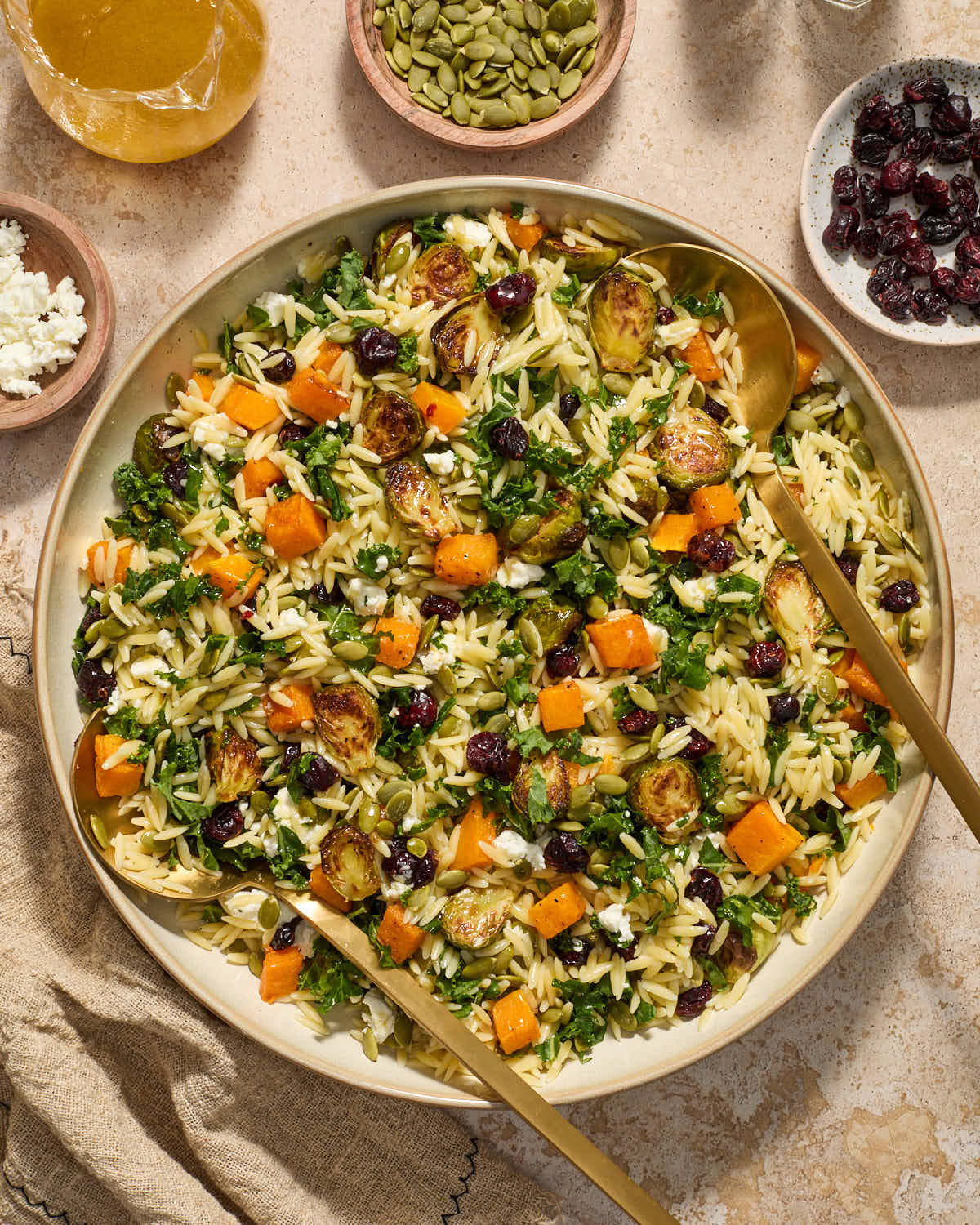 Fall orzo salad in a large shallow bowl with serving spoons inserted into it and toppings surrounding it.