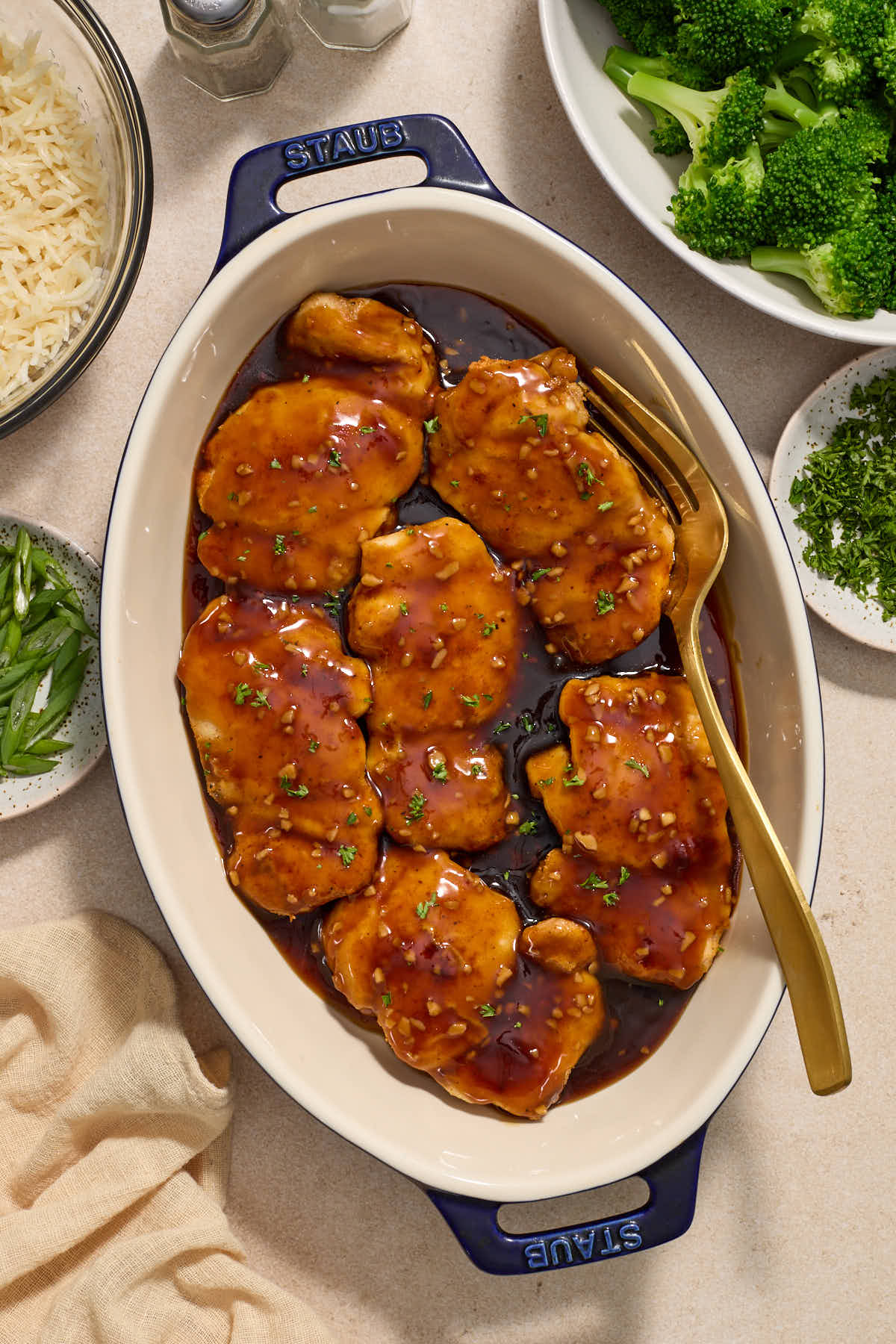 Chicken thighs covered in sauce in an oval dish with garnishes, broccoli and rice on the side.
