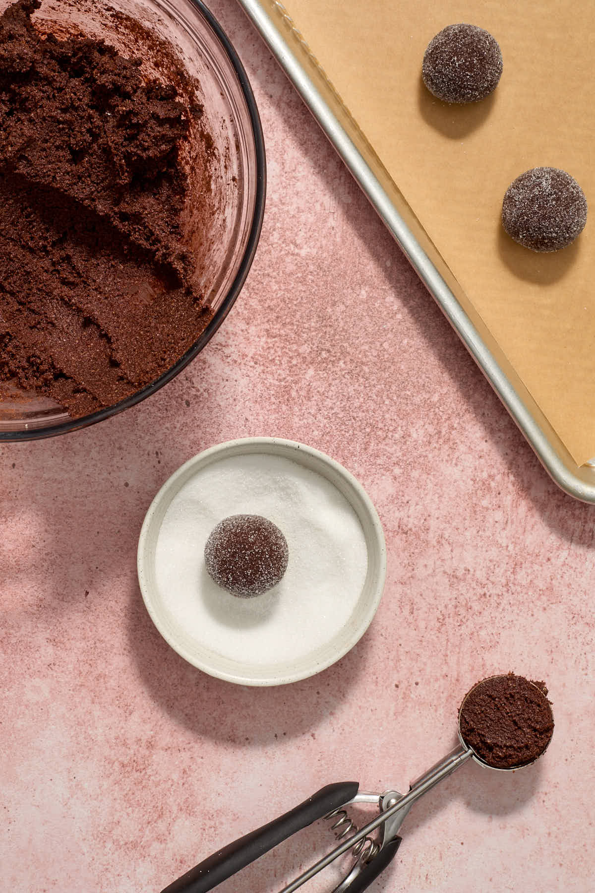 Dough rolled into a ball and rolled in a bowl of sugar with the bowl of dough and a baking sheet on the side.