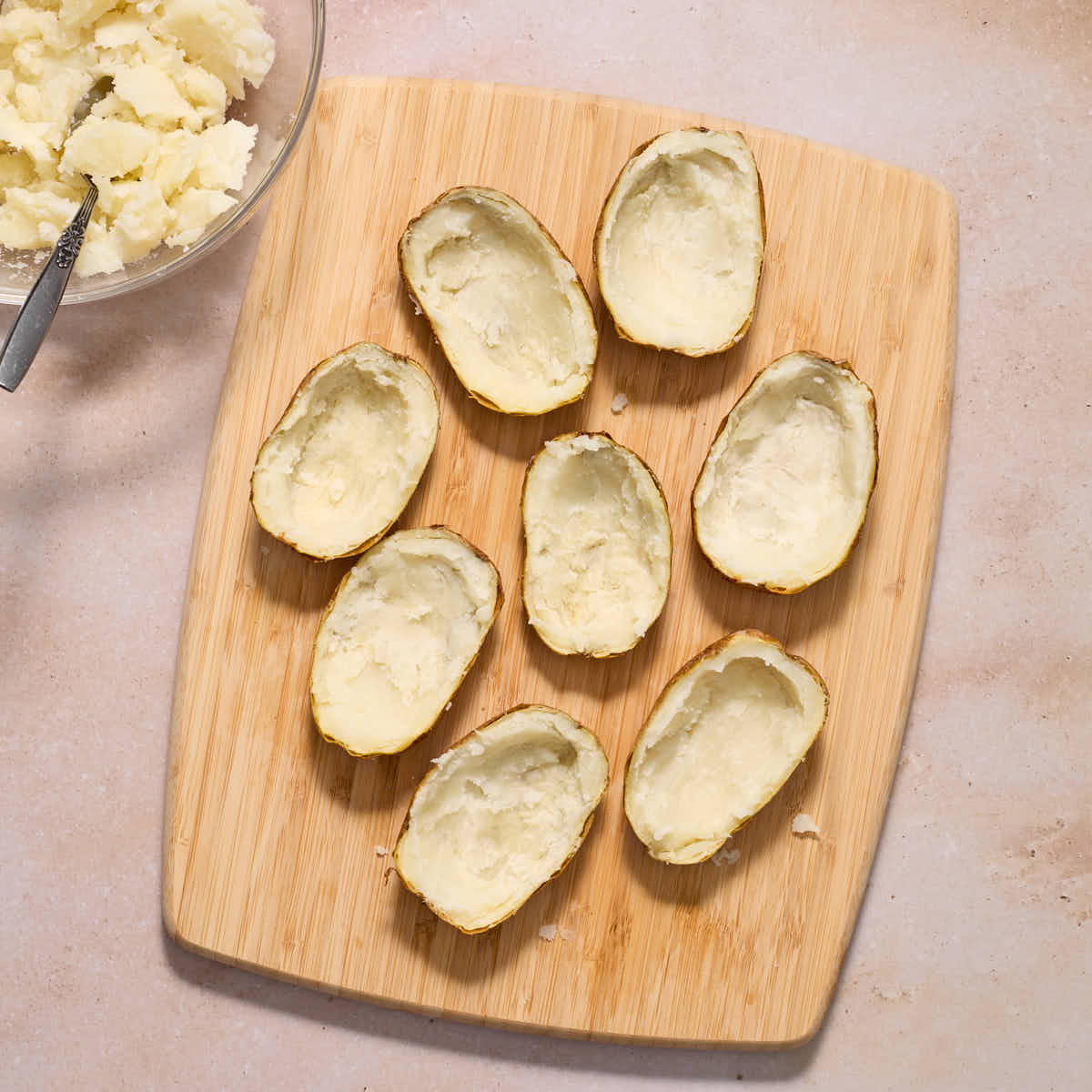 Baked potatoes cut in half horizontally on a wooden cutting board with the flesh scooped out into a glass bowl.