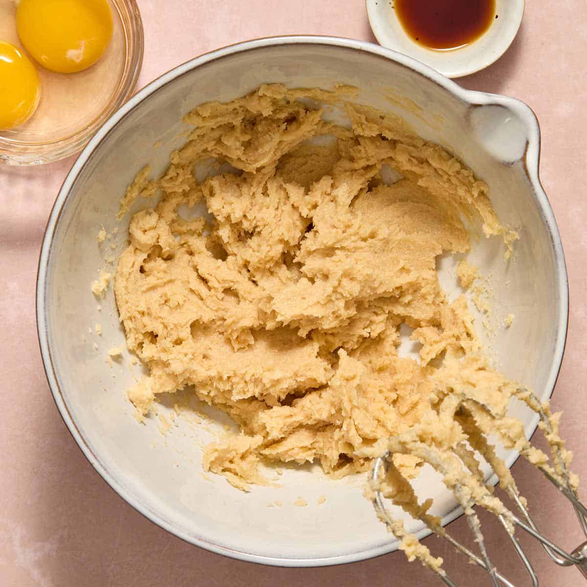 Butter and sugars creamed together in a white bowl with eggs and vanilla on the side.