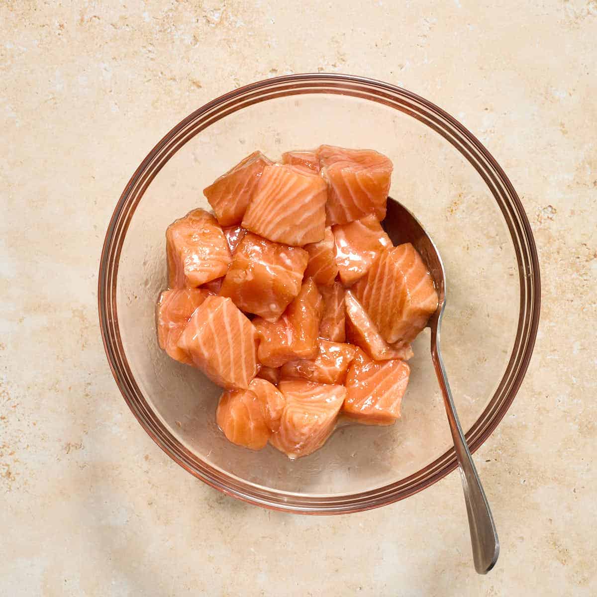 Raw salmon cubes in a glass bowl and coated with oil and soy sauce.