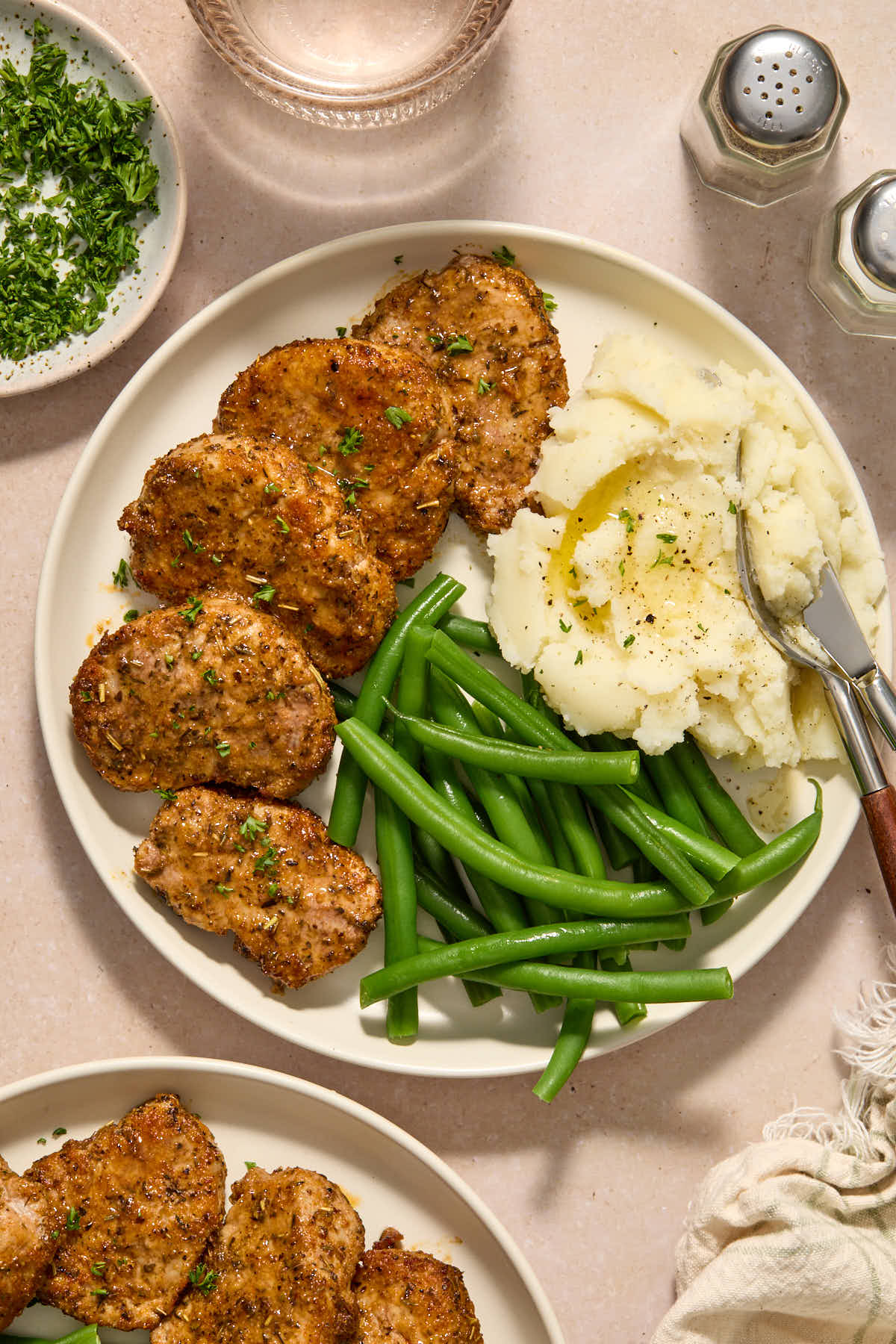 Overhead of pork served on plates with green beens and mashed potatoes with chopped parsley on the side.