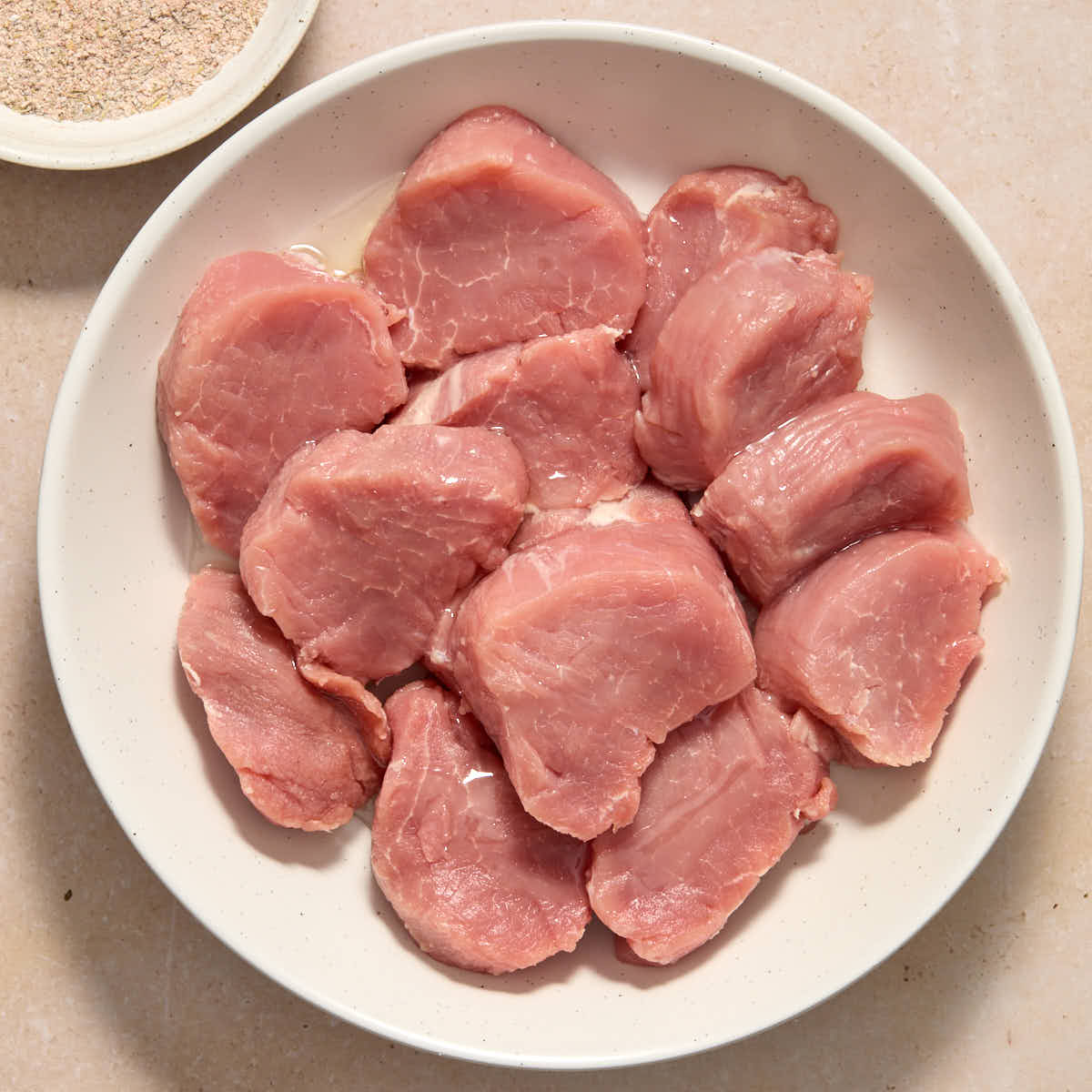 Sliced pork tenderloin in a white bowl, coated in oil, with seasoning on the side.
