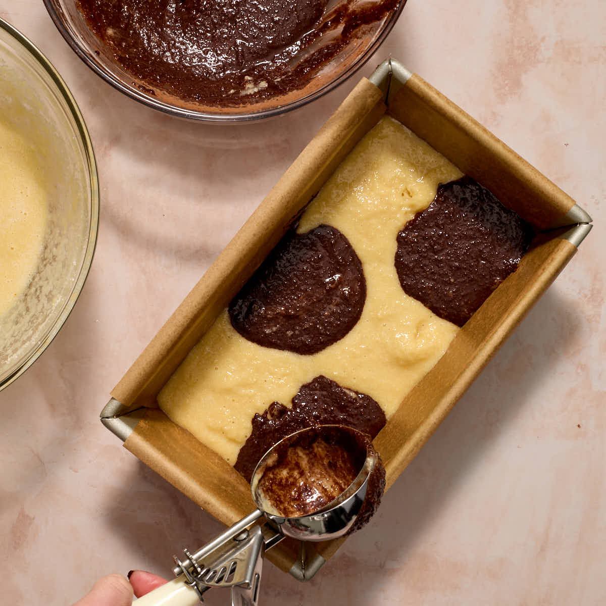 Vanilla and chocolate batters being scooped into a loaf pan in an alternating pattern.