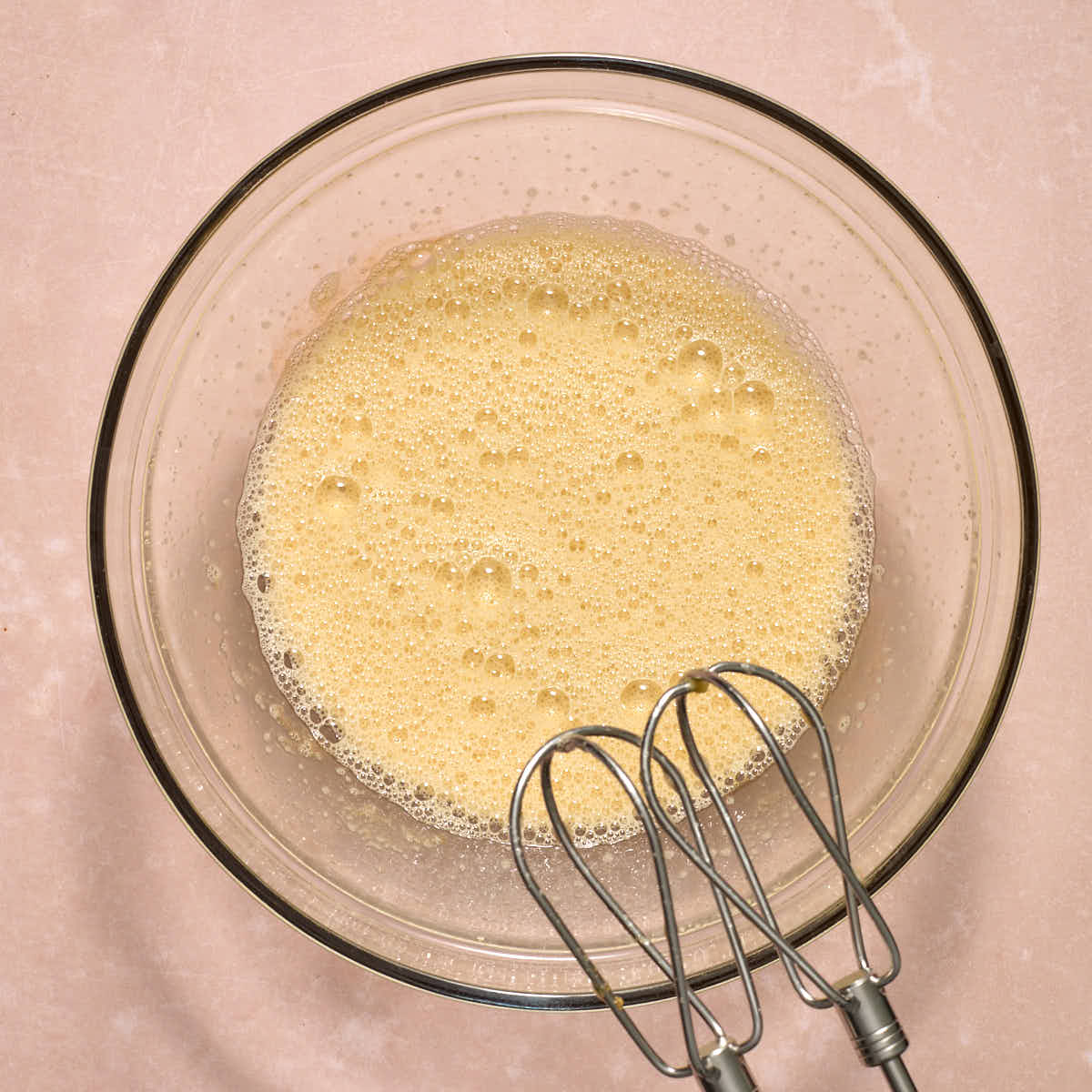 Eggs and brown sugar beaten together in a glass bowl with bubbles formed on top.