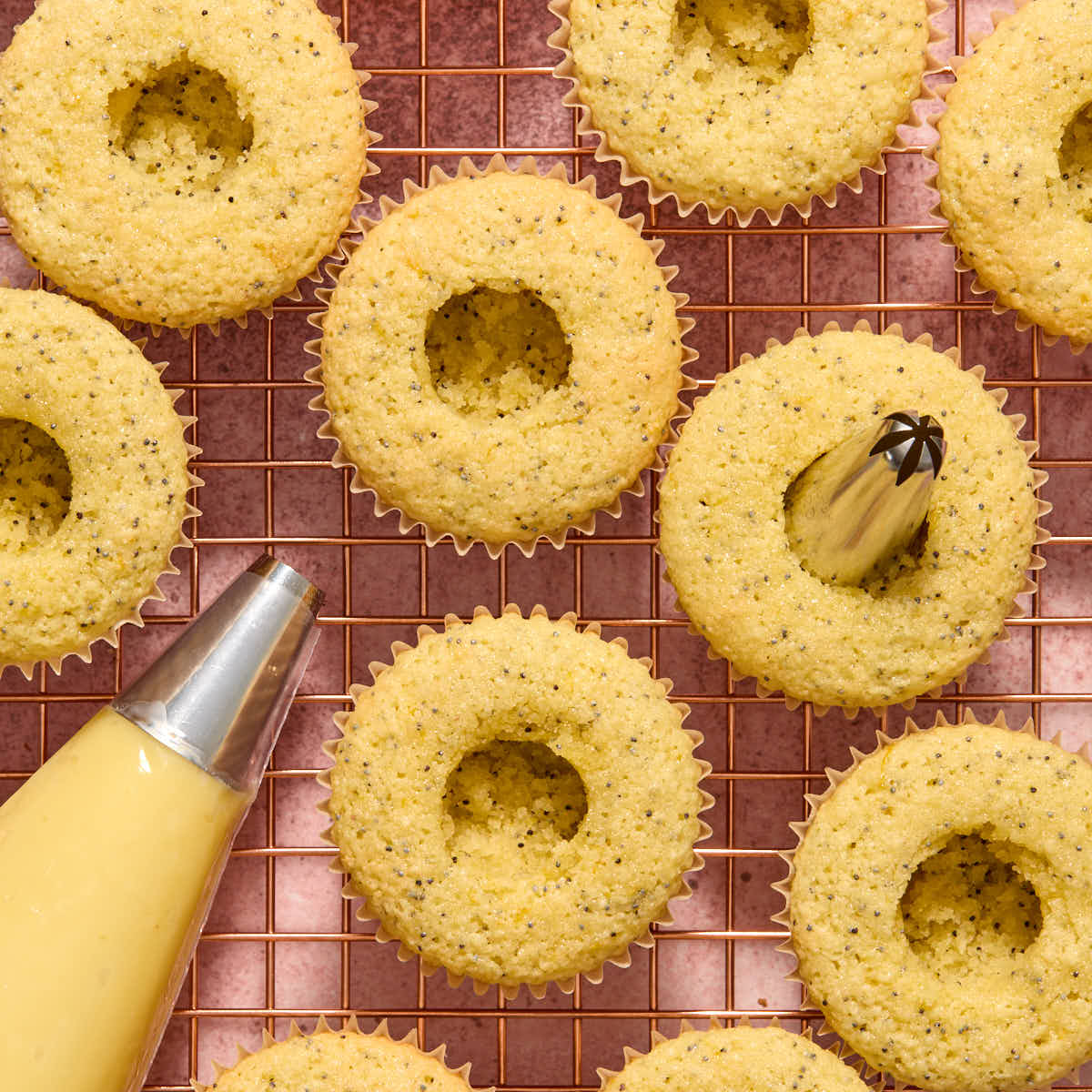 Cupcakes on a wire rack with center holes carved out and lemon curd in a piping bag.