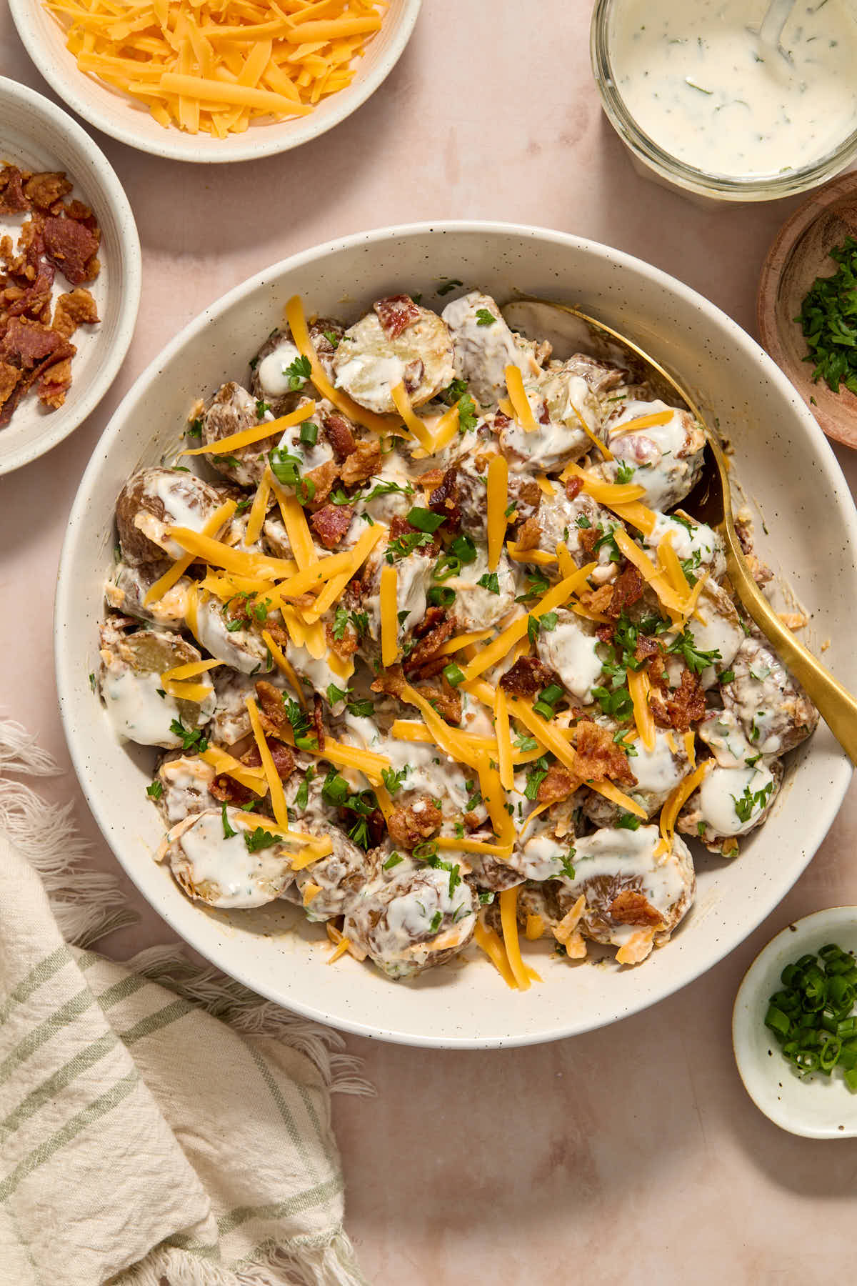 Potato salad in a white bowl with a spoon and extra cheese, bacon, dressing and chives on the side.