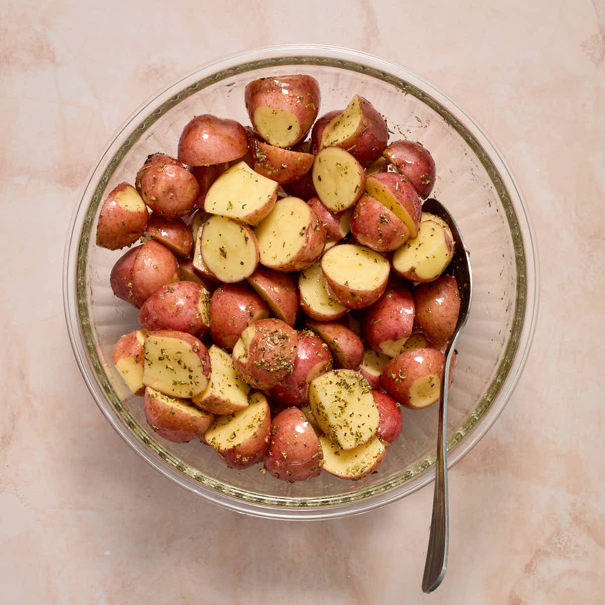 Potatoes tossed with oil and seasoning in a glass bowl with a spoon inserted into it.