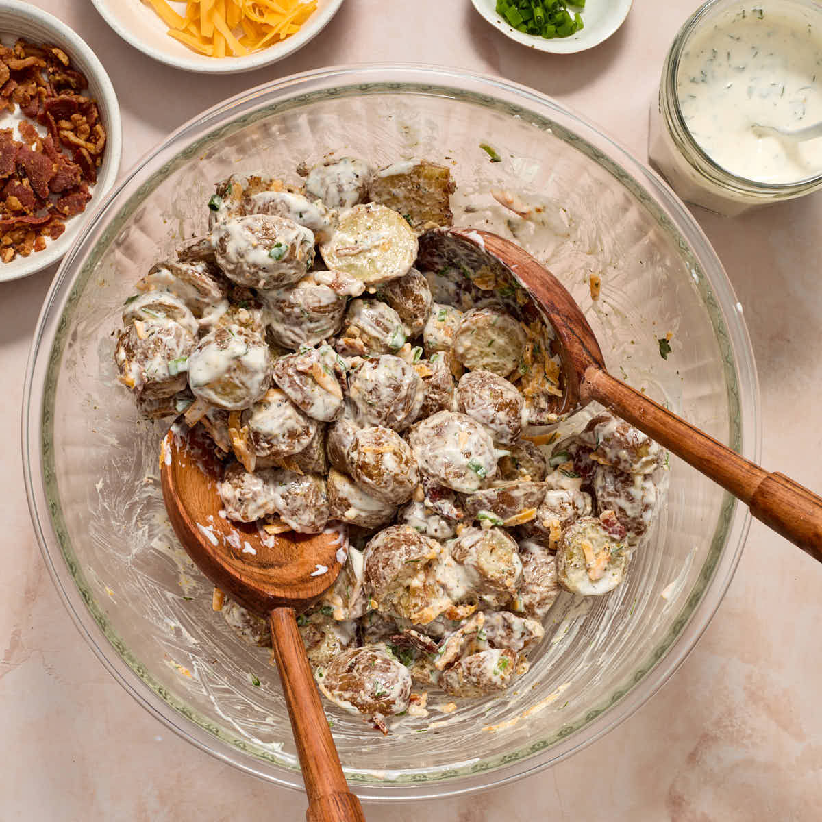Potatoes mixed with cheese, bacon, chives and dressing in a glass bowl with wooden spoons.