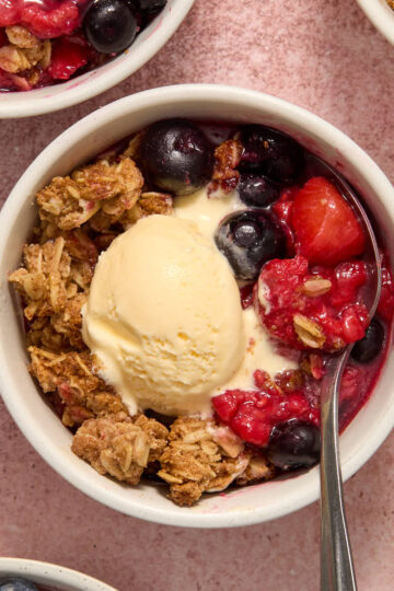 Air fryer berry crisp in a white ramekin with a spoon inserted and a scoop of ice cream on top.