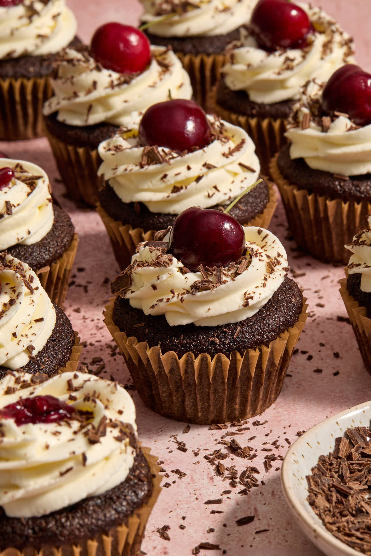 Black forest cupcakes arranged on a pink surface with chocolate shavings scattered around.