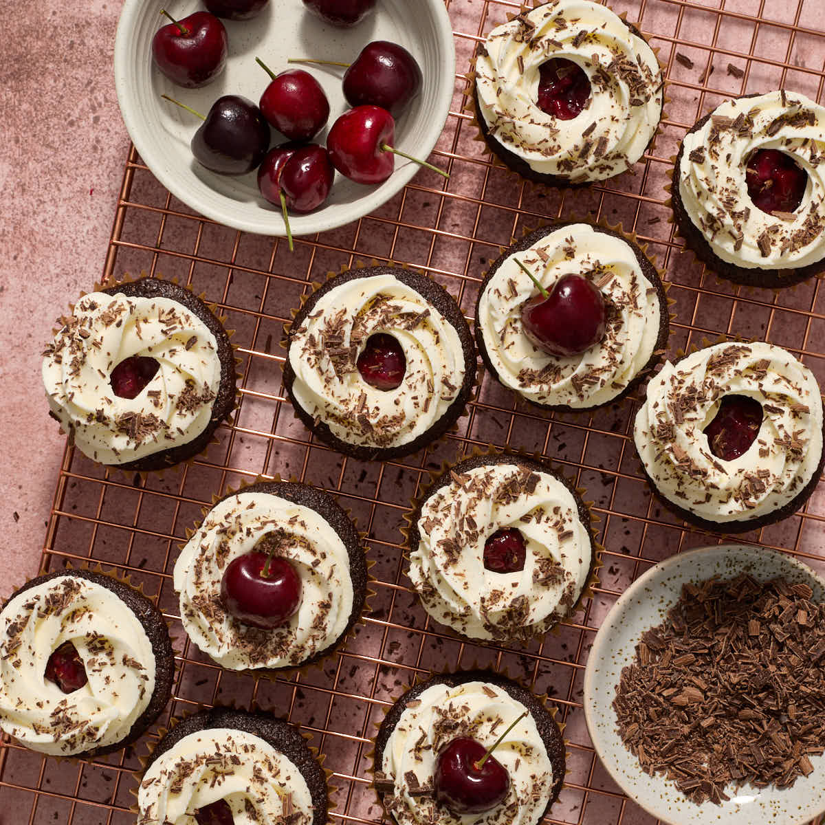 Frosted cupcakes on a wire rack, topped with chocolate shavings and cherries.