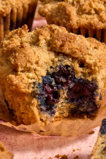 Almond flour blueberry coffee cake muffin with a bite removed and resting on a parchment muffin liner.