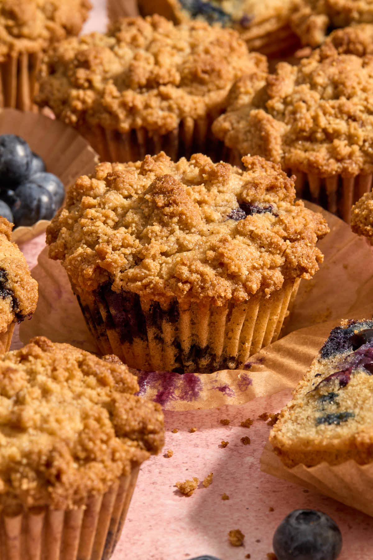 Blueberry coffee cake muffin on parchment wrapper with other  muffins and blueberries around it.