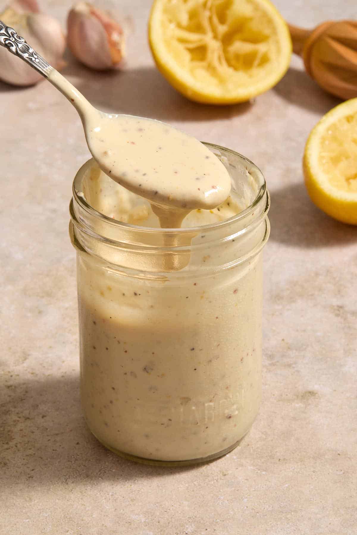 Dressing in a glass jar with a spoon in it and lemons and garlic in the background.