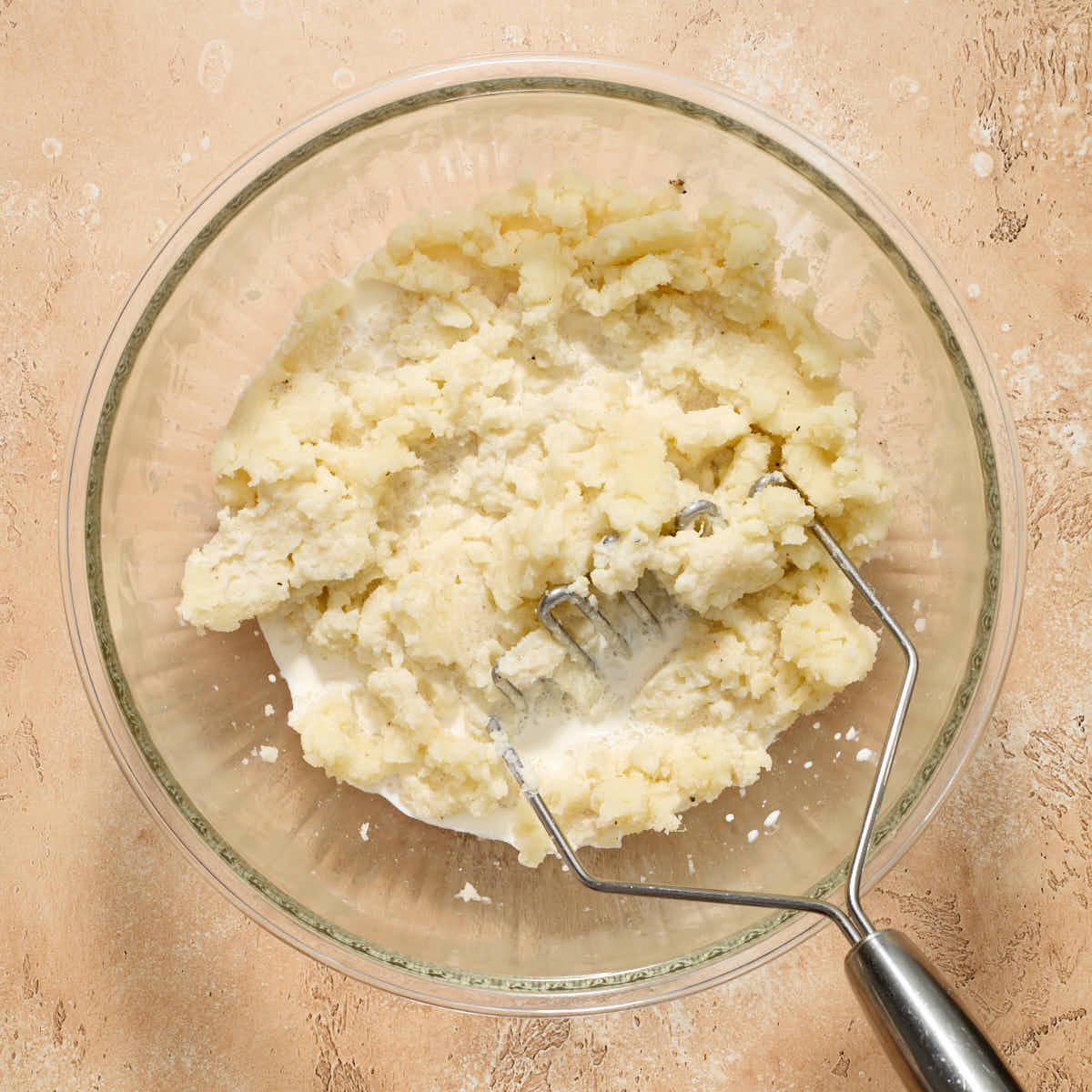 Cream added to the bowl of mashed potatoes with the potato masher in the bowl.