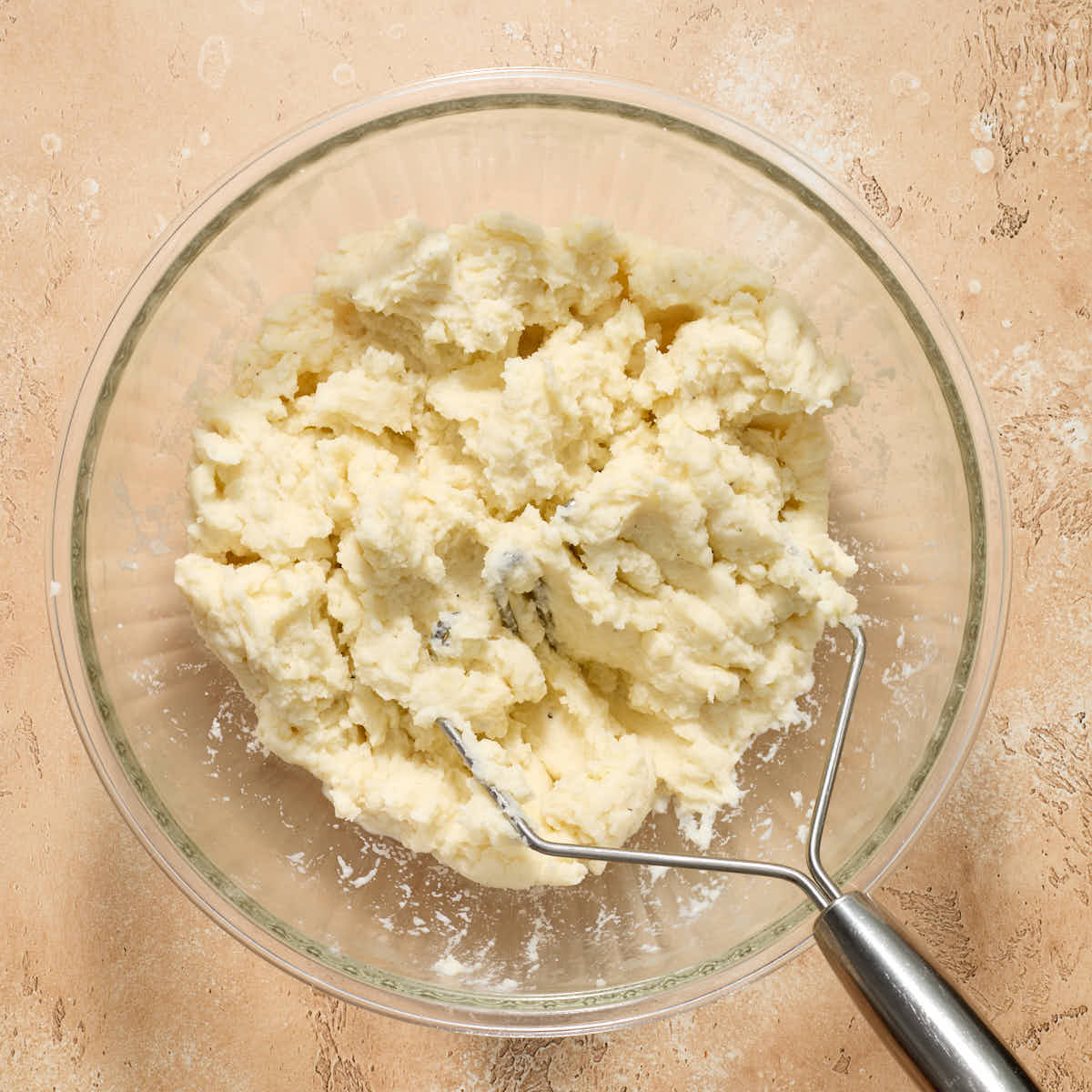 Fully mashed potatoes in a glass bowl with the potato masher.