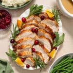 Air fryer turkey breast sliced and served on a platter with fresh herbs, cranberries and orange wedges.