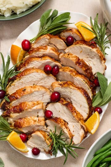 Air fryer turkey breast sliced and served on a platter with fresh herbs, cranberries and orange wedges.