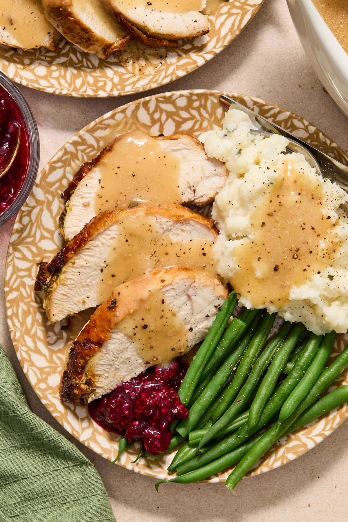 Slices of turkey served on a plate with mashed potatoes, green beans, cranberry sauce and gravy.